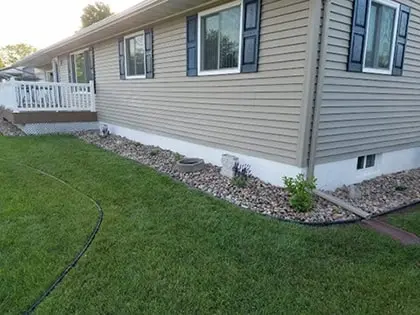decorrock-420pxw Single-story beige house with blue shutters, surrounded by a neatly landscaped yard. A rock border with sparse plants lines the house, and the lawn is green and well-maintained. A black hose runs across the grass.