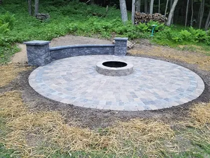 firepit-420pxw A circular stone patio with a built-in fire pit in the center and a curved stone bench at the back, set in a grassy outdoor area near trees.