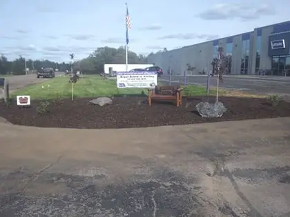 mulch-420pxw A landscaped area with mulch, a bench, two large rocks, and a sign in the background near a large commercial or industrial building with a parking lot and vehicles. An American flag is visible on a pole.