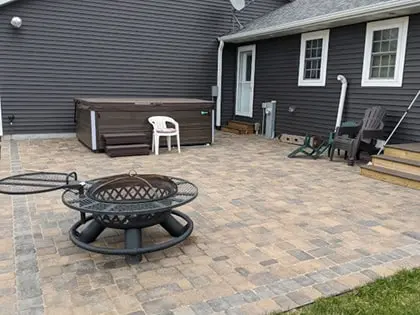 patio-420pxw A patio with stone pavers features a round metal fire pit, a hot tub, plastic chairs, and Adirondack chairs next to a dark gray house with white trim and wooden steps leading to a door.