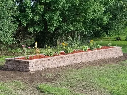 planter-420pxw A long rectangular raised garden bed made of stacked stones, filled with mulch and various small flowering plants, sits on a grassy lawn with dense green trees in the background.