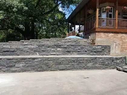 wall2-420pxw A multi-level gray stone retaining wall stands in front of a red brick building with a wooden porch and railing, surrounded by trees and greenery.